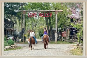 Two Girls riding horses at the entrance sign of Love Valley NC (Love Valley is Written in Red between 2 wooden posts.