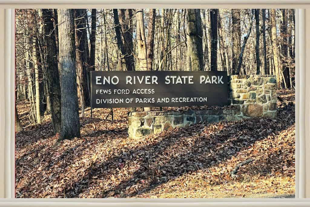 The Eno River State Park Fews Ford Access sign (brown with white letters ona stone support wall. Trees are bare and brown leaves on the ground in winter.  

History at Eno River State Park