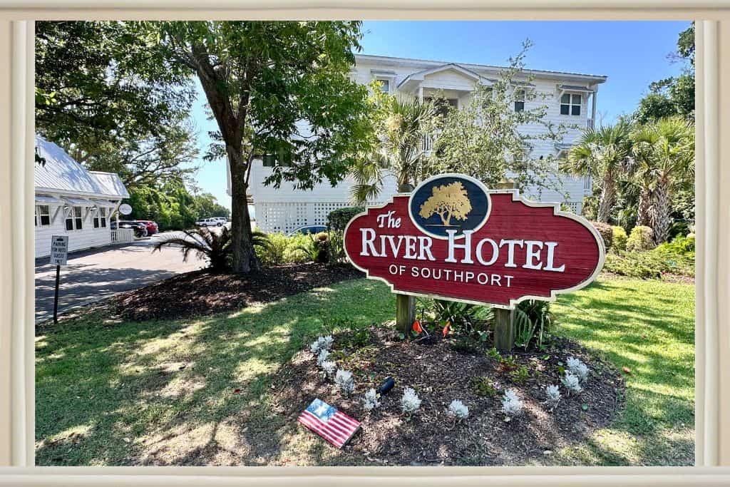 red sign that says river hotel and a navy circole top middle with a gold tree. behind the sign is greenery and the whilte building of the River hotel behind the trees. Place to stay in Southport